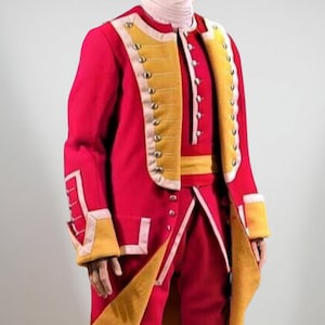 Peut inclure: Un costume historique rouge et or, comprenant un long manteau avec des garnitures et des boutons dorés, un pantalon bouffant assorti et des chaussures à boucle noires. La tenue comprend une chemise à col blanc et une ceinture jaune. Costume de reconstitution historique.