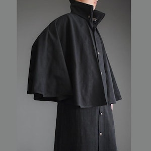 May include: A black cloak and coat combination. The cloak has a high collar and a cape-like design over the shoulders. The coat underneath features a button-down front and a knee-length hem. The fabric appears to be a textured, dark material.