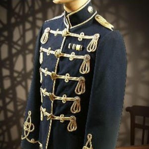 Könnte beinhalten: Marineblaue Jacke im Militärstil mit goldfarbenen Seil- und Knopfdetaills. Die Jacke hat einen hohen Kragen und Schulterklappen und wird auf einer Schaufensterpuppe präsentiert. Die Jacke hat dekorative goldene Verzierungen.