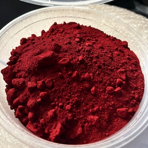 May include: A clear plastic container filled with a vibrant, deep red powder. The powder has a fine, granular texture with small, irregularly shaped chunks throughout. The container is partially open, revealing the contents.