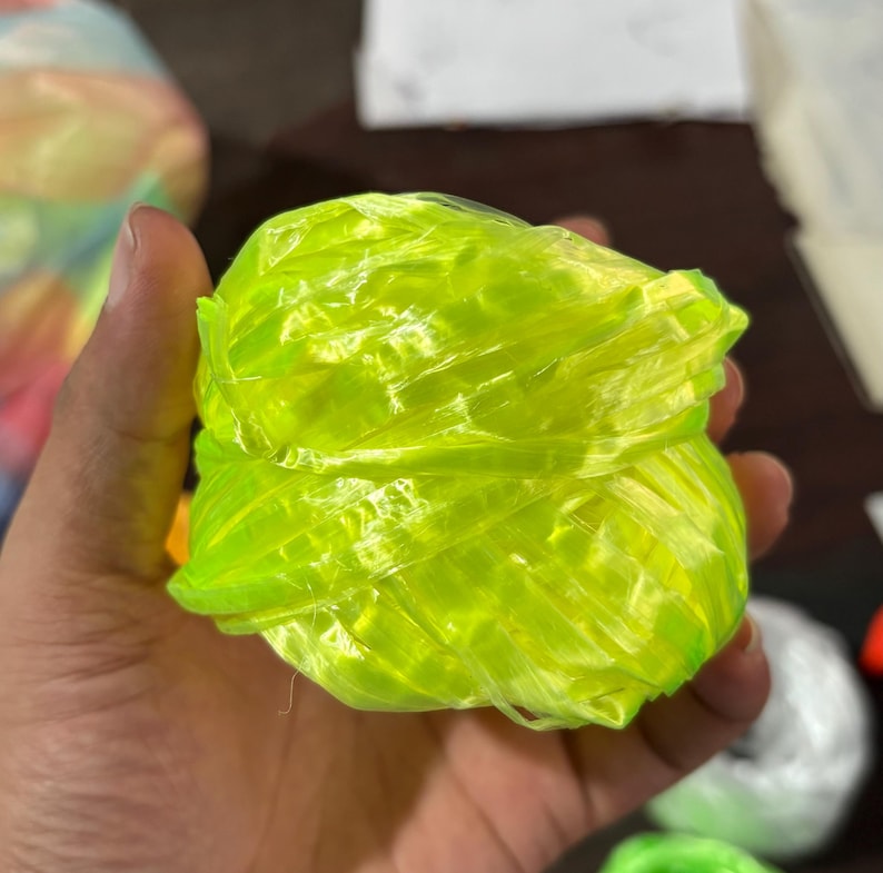 May include: A ball of bright neon yellow twine, held in a hand. The twine appears to be made of a synthetic material, with a slightly translucent quality. The twine is tightly wound into a compact ball shape.