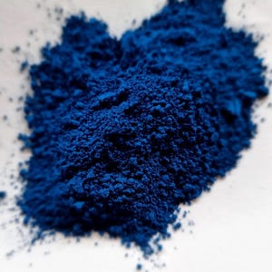 Puede incluir: Primer plano de un mont&oacute;n de polvo azul intenso y vibrante. El polvo tiene una textura fina y granulada y est&aacute; densamente empaquetado, creando un color rico y saturado. El polvo est&aacute; sobre una superficie blanca.