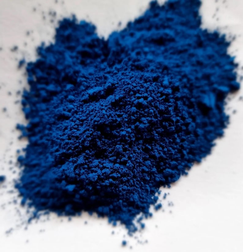 Puede incluir: Primer plano de un mont&oacute;n de polvo azul intenso y vibrante. El polvo tiene una textura fina y granulada y est&aacute; densamente empaquetado, creando un color rico y saturado. El polvo est&aacute; sobre una superficie blanca.