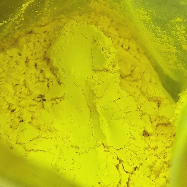 Puede incluir: Primer plano de un polvo amarillo brillante, posiblemente pigmento o tinte, que llena el encuadre. El polvo tiene una apariencia texturizada y granulada con grumos y grietas visibles. El color es uniforme.
