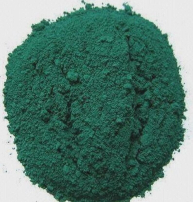 Puede incluir: Un mont&oacute;n de polvo finamente molido, de color verde azulado oscuro. El polvo tiene un aspecto texturizado y granulado, con variaciones de tono que crean luces y sombras. La forma general es un mont&iacute;culo redondeado sobre un fondo blanco.