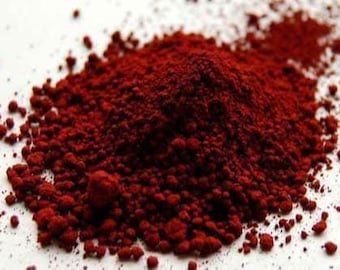 May include: A close-up of a pile of deep red powder with a granular texture. The powder is scattered on a white surface, with small granules surrounding the main pile. The color is consistent throughout.