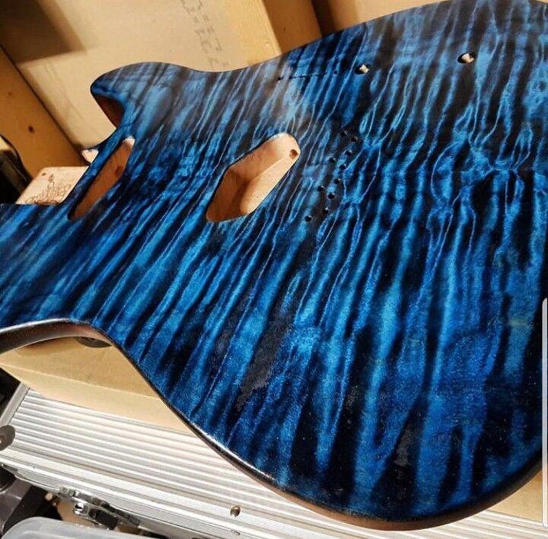 May include: A partially constructed electric guitar body with a vibrant blue flame maple finish. The wood grain creates a flowing pattern. The body has cutouts for pickups and other hardware, showcasing the instrument's design.