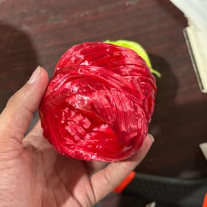 May include: A tightly wound ball of bright red raffia, held in a hand. The raffia appears to be made of a shiny, slightly translucent material. A small portion of green raffia is visible in the background.