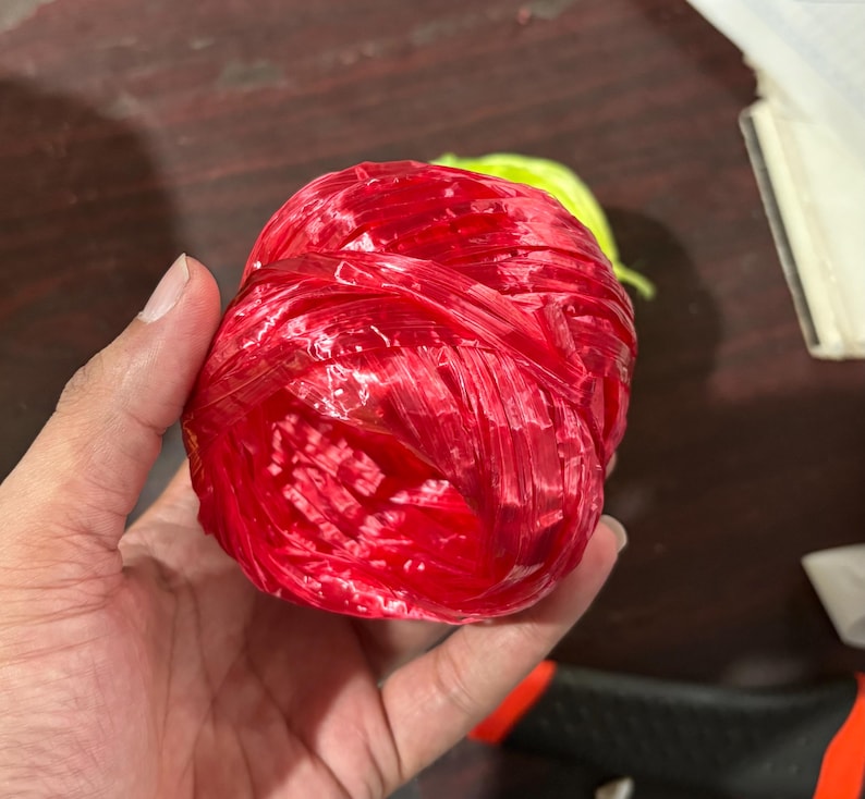 May include: A tightly wound ball of bright red raffia, held in a hand. The raffia appears to be made of a shiny, slightly translucent material. A small portion of green raffia is visible in the background.
