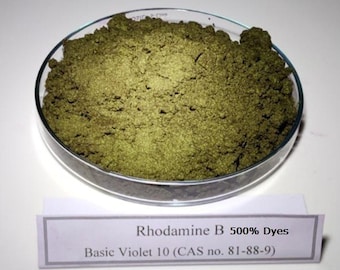 May include: A clear glass petri dish filled with a coarse, olive-green powder. Below the dish, a white rectangular label displays the text: Rhodamine B 500% Dyes, Basic Violet 10 (CAS no. 81-88-9). The substance has a granular, matte texture.