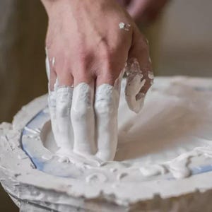 Könnte beinhalten: Nahaufnahme einer Hand, die mit weißem Material bedeckt ist und in eine weiße Form drückt. Die Form hat einen blauen Rand und ist von einer rauen, strukturierten Oberfläche umgeben. Das Bild deutet auf einen handwerklichen oder künstlerischen Prozess hin.