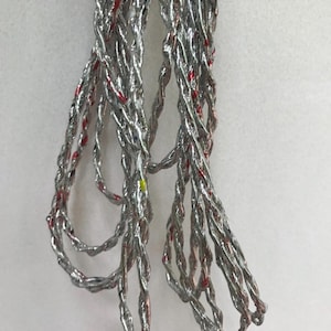 May include: Close-up of several strands of silver-colored, twisted wire. The wire has red and yellow accents throughout. The strands are hanging against a white background.