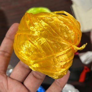 May include: A ball of bright yellow plastic twine, tightly wound and slightly translucent. The twine has a shiny appearance and is held in a hand, with other objects blurred in the background.