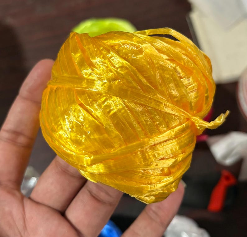 May include: A ball of bright yellow plastic twine, tightly wound and slightly translucent. The twine has a shiny appearance and is held in a hand, with other objects blurred in the background.