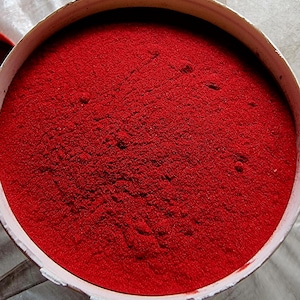 May include: A top-down view of a round, white container filled with a vibrant red powder. The powder has a fine, textured appearance, filling the container to the brim. The container's rim is visible, contrasting with the intense color of the powder.