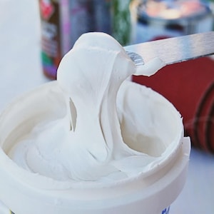May include: A close-up of a white, creamy substance being scooped from a white container with a metal spatula. A roll of red sandpaper is in the background, along with other blurred items. The substance appears to be a filler or sealant.