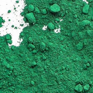 Può includere: Una vibrante polvere di pigmento verde smeraldo con una consistenza fine e granulosa. La polvere è densamente compatta con piccoli grumi irregolari sparsi sulla superficie, creando un profilo cromatico ricco e saturo con ombre profonde.