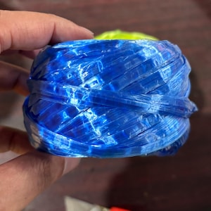May include: A tightly wound ball of blue plastic twine, held in a hand. The twine has a woven texture and a glossy finish, with variations in the blue color. The twine is likely used for crafting or packaging.