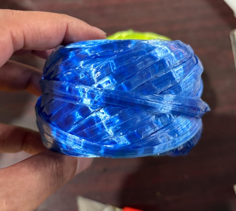 May include: A tightly wound ball of blue plastic twine, held in a hand. The twine has a woven texture and a glossy finish, with variations in the blue color. The twine is likely used for crafting or packaging.