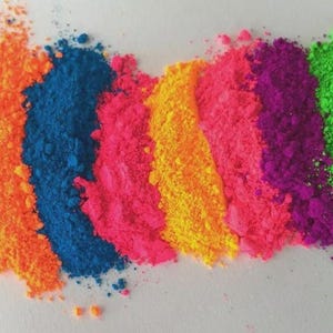 Puede incluir: Una imagen vibrante que muestra un espectro de polvos de colores. Los polvos están dispuestos en líneas paralelas, mostrando tonos de amarillo neón, naranja, azul, rosa, amarillo, morado y verde. Los polvos tienen una textura fina y granular.