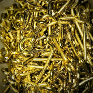 May include: A close-up shot showcases a pile of shiny, golden-colored screw eye hooks. The hooks are densely packed, with their eyelets and screw threads clearly visible. The overall impression is of a collection of hardware components.