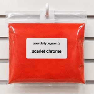 Può includere: Un sacchetto di plastica trasparente pieno di pigmento rosso brillante, etichettato "yourdailypigments" e "scarlet chrome" su un rettangolo bianco. Il sacchetto è appeso a un gancio bianco su uno sfondo bianco scanalato.