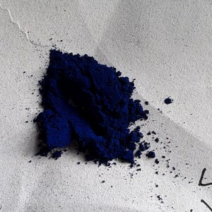 以下が含まれることがあります： 濃い青色の粉末の山が、質感のあるオフホワイトの表面に置かれています。 粉末は狭い範囲に集中しており、いくつかの粒子が散らばっています。 表面にはわずかな折り目があり、右下隅には手書きの数字があります。