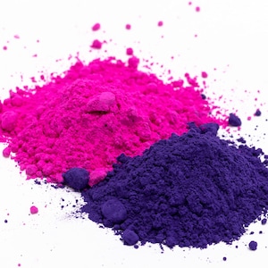Puede incluir: Primer plano de dos montones de pigmentos en polvo de colores. Uno es de un magenta brillante, el otro de un &iacute;ndigo profundo. Los polvos est&aacute;n finamente molidos y parecen suaves, con gr&aacute;nulos dispersos en los bordes, sobre un fondo blanco.
