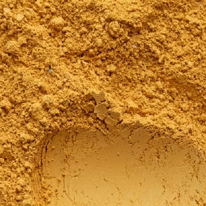 Può includere: Una polvere fine e secca in una vibrante tonalità giallo senape. La consistenza è irregolare, caratterizzata da piccoli grumi e una grana granulosa su tutta la superficie. Il materiale appare sciolto e opaco, con una saturazione di colore densa.