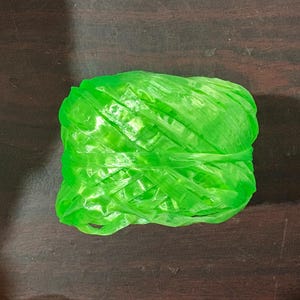 May include: A tightly wound ball of bright green plastic twine, resting on a dark wooden surface. The twine appears to be made of thin, flat strands, tightly wrapped together. The overall shape is rectangular.