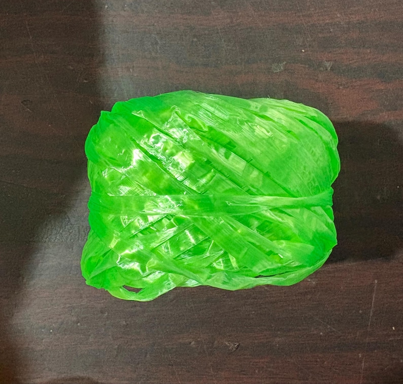 May include: A tightly wound ball of bright green plastic twine, resting on a dark wooden surface. The twine appears to be made of thin, flat strands, tightly wrapped together. The overall shape is rectangular.