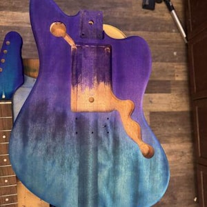 May include: A partially assembled guitar body with a gradient finish in shades of blue and purple. The body has cutouts for the pickups, bridge, and neck. The neck and headstock are also visible, with a blue finish.