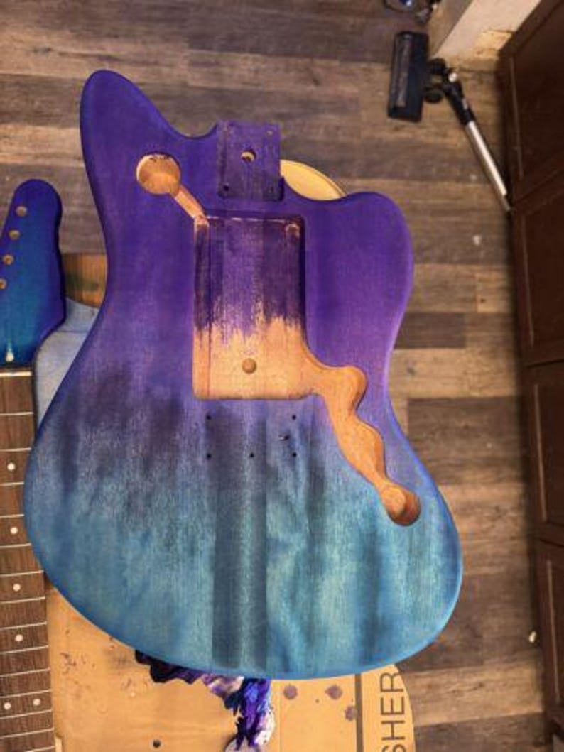 May include: A partially assembled guitar body with a gradient finish in shades of blue and purple. The body has cutouts for the pickups, bridge, and neck. The neck and headstock are also visible, with a blue finish.