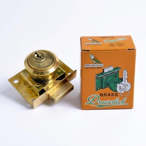 Könnte beinhalten: Ein Messing-Schubladenschloss mit einem runden Schlüsselloch und einer rechteckigen Montageplatte, neben der originalen orangefarbenen Pappschachtel. Die Schachtel trägt die Aufschrift "BRASS Drawerlock", "No. 0501", "ONE ONLY" und "WREN BIRD BRAND".