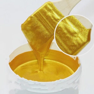 May include: A close-up of a white container filled with shimmering gold paint. A wooden spatula holds a large amount of the paint, with some dripping into the container. A circular inset shows a magnified view of the paint's texture.