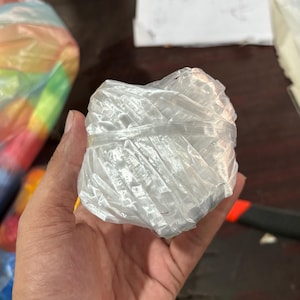 May include: A tightly wound ball of clear, shiny twine. The twine is held in a hand, with the ball appearing to be about 3 inches in diameter. The twine is made of a synthetic material and is likely used for crafting or packaging.
