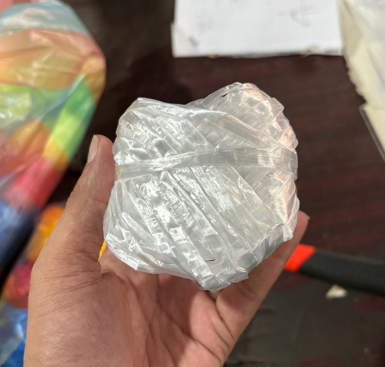 May include: A tightly wound ball of clear, shiny twine. The twine is held in a hand, with the ball appearing to be about 3 inches in diameter. The twine is made of a synthetic material and is likely used for crafting or packaging.