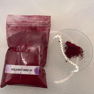 Könnte beinhalten: Ein durchsichtiger Plastikbeutel gefüllt mit tiefrotem Pulver, beschriftet mit "SOLVENT RED 41". Eine kleine Menge des gleichen roten Pulvers befindet sich auf einer klaren, runden Glasschale. Das Pulver scheint ein Pigment oder Farbstoff zu sein.