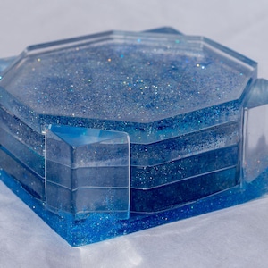 May include: A set of four blue resin coasters with a glittery, ocean-like design. The coasters are hexagonal and sit in a matching holder. The resin has varying shades of blue, creating a layered effect. The holder has a rectangular shape.