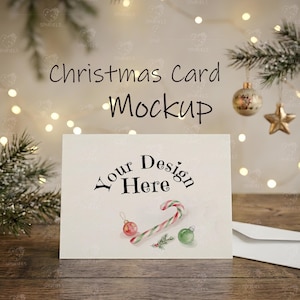 May include: A white folded Christmas card mockup featuring the text Your Design Here in black, with a watercolor illustration of a candy cane, a red ornament, a green ornament, and pine needles. A white envelope sits to the right.