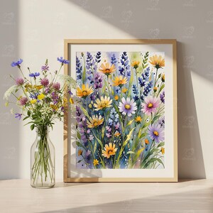 May include: A framed watercolor print of wildflowers in shades of yellow, purple, and pink. A clear glass vase with a bouquet of colorful wildflowers sits to the left of the framed print. The frame is a light wood color.
