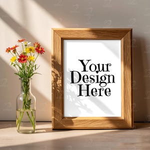 May include: A wooden picture frame with the text "Your Design Here" on a white background. A small glass vase with colorful flowers sits to the left of the frame. The frame is made of light brown wood.