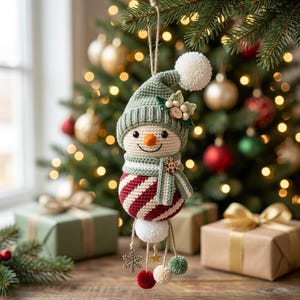 May include: A crocheted snowman ornament featuring a sage green hat with a white pom-pom, a striped scarf with a wooden snowflake accent, and a red and white striped body. It includes dangling charms of a snowflake, a star, and small pom-poms.
