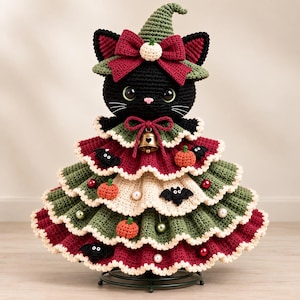 May include: A crocheted black cat figure wearing a green pointed hat with a red bow and a small white pumpkin accent. The body is a tiered, ruffled dress in alternating layers of red, green, and cream, decorated with small pumpkins, bats, and beads.