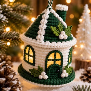 Könnte beinhalten: Ein gehäkeltes Haus-Ornament aus weißem und dunkelgrünem Garn. Die Struktur verfügt über ein kegelförmiges Dach, einen Schornstein, bogenförmige Fenster und eine zentrale Tür. Dekorative grüne Blätter und weiße Beeren zieren Dach und Basis.