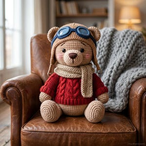 Op de afbeelding: Een gehaakte teddybeer met een beige vliegeniersmuts met blauwe bril, een beige gebreide sjaal en een rode kabeltrui. De beer heeft lichtbruine garenvacht, zwarte ogen, een donkerbruine geborduurde neus en roze blosjes op de wangen.