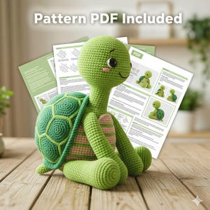 Pode incluir: Uma tartaruga de crochê verde e verde claro, feita à mão, com um rosto sorridente e uma barriga listrada. Está sentada numa superfície de madeira, com instruções de padrão no fundo. O texto "Pattern PDF Included" é visível.