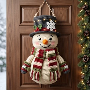 May include: A crocheted snowman wall hanging featuring a dark gray top hat with a white snowflake, green holly, and red berries. The snowman has a carrot nose, pink cheeks, and a striped red, white, and green scarf with matching mittens.