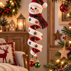 May include: A crocheted snowman ornament featuring a red and white Santa hat, a striped red and green scarf, and a long white spiral body decorated with green and red snowflake patterns. The ornament ends with two shiny red and green ball baubles.