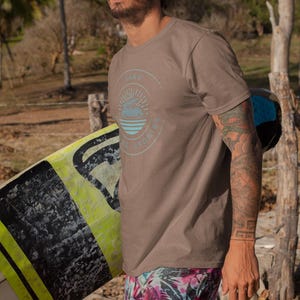 May include: A man in sunglasses wearing a brown t-shirt with a circular graphic and the words "Sand Stories" in light blue. He is wearing colourful shorts and holding a surfboard on a beach.